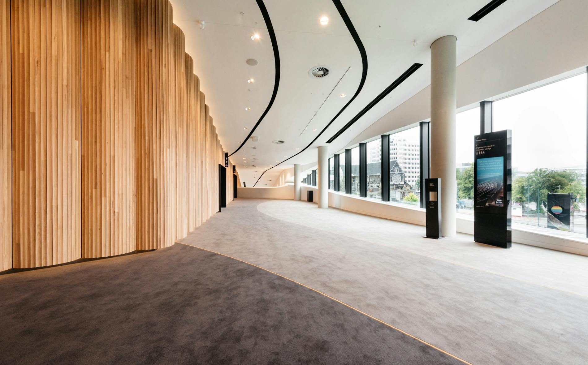 A smooth, curving corridor features a wooden wall and plush carpet. Te Pae branding is visible, with large windows showcasing an urban landscape outside, highlighting the modern interior design.